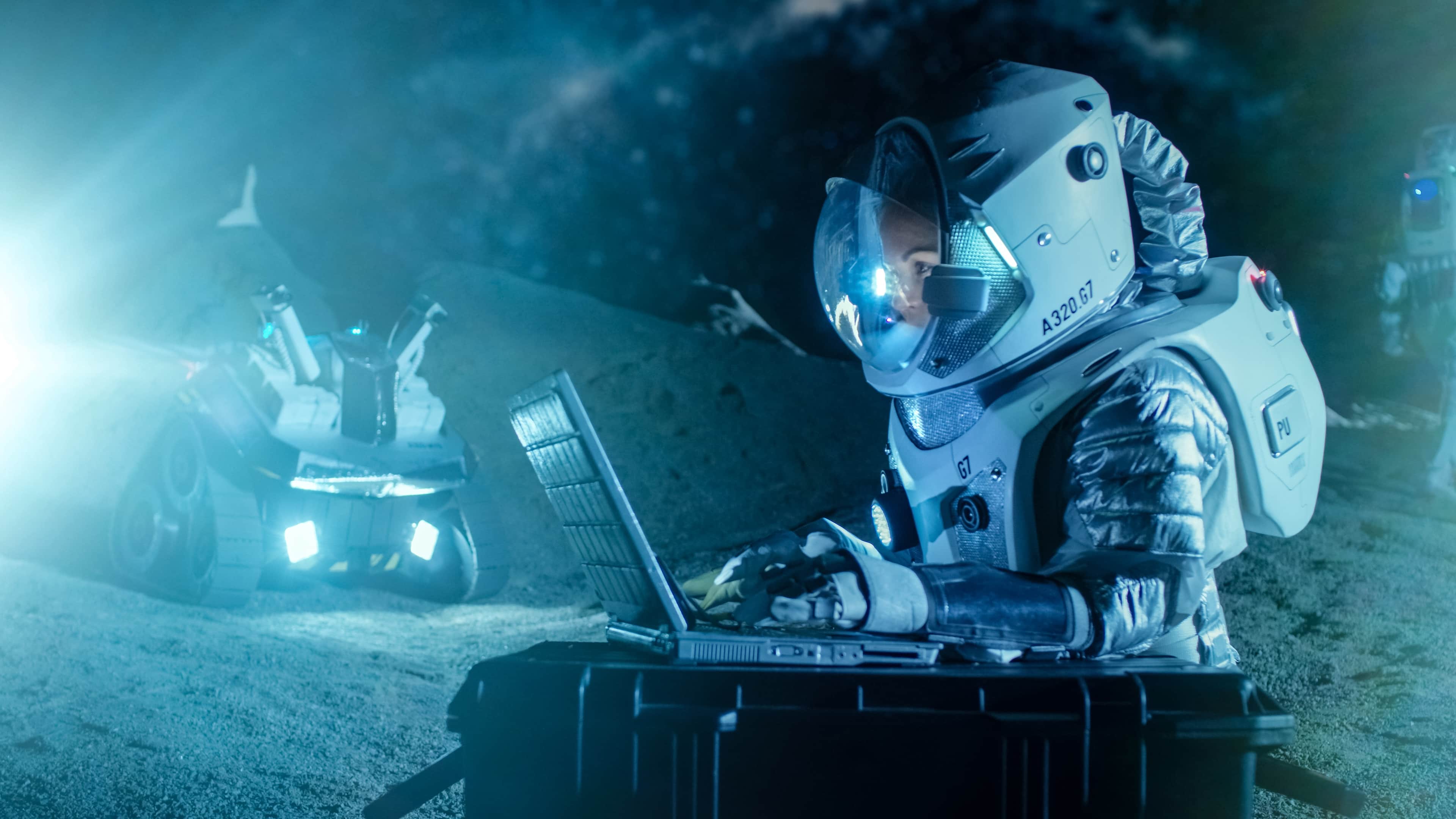Female Astronaut Wearing Space Suit Works on a Laptop, Exploring Newly Discovered Planet, Communicating with the Earth. In the Background Her Crew Member and Space Habitat. Colonization Concept.